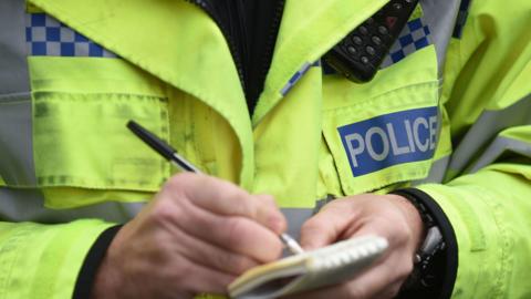 Close up shot of Police officer taking notes on to a notepad. He is wearing a fluorescent jacket and his face is not shown.