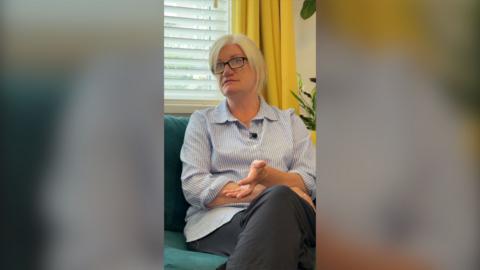 Blonde woman wearing a light blue stripped shirt sits on sofa during interview