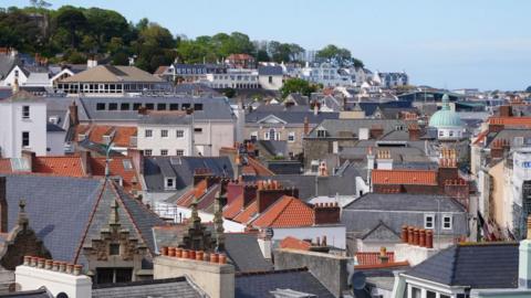 Am image of houses and red and grey rooftops in Guernsey, with trees in the background. The sky is blue.
