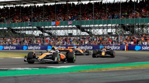 Formula 1 cars racing during the British Grand Prix at Silverstone. Cars from McLaren, Red Bull and Ferrari are all shown against the backdrop of a packed grandstand.
