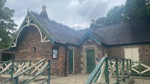 A red brick, single-storey Victorian pavilion with a slanted roof with green wooden trim around it. There are two green doors on the right of the building and a green and cream wooden banister around the path in front of it.  The paintwork is chipped, making the building and banisters look run down. There is a bulletins board at the front of the building.