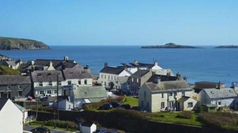 A view of houses against a backdrop of the sea. A set of houses, some detached.  A cliff head can be seen in the background.