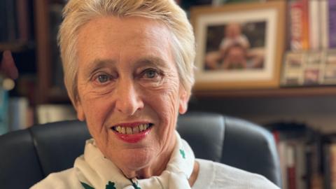 Anne Holland has short white hair and is wearing a white scarf and top. She is looking at the camera and smiling. There are books and a framed photo on a shelf behind her.