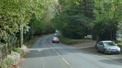 Cars travel along a road. Trees line either side of the carriageway.
