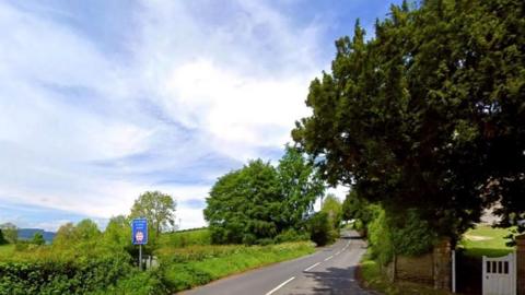 A country road snaking up a hill, with greenery to the left and right. There are hills in the distance on the left and a white gate bottom right of the image.