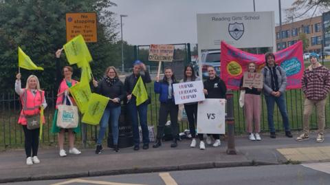 Ten people standing outside the gates of a school holding flags and placards.