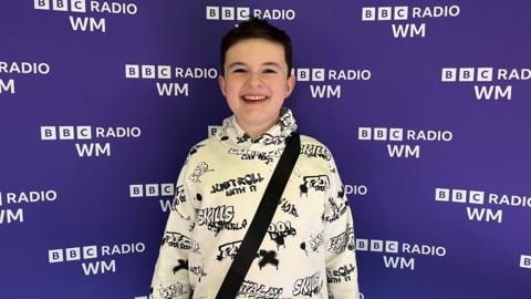 Finley Hill, a 12-year-old boy, is stood against a purple BBC Radio WM banner. He is wearing a white hoodie with black graffiti-style writing on it.