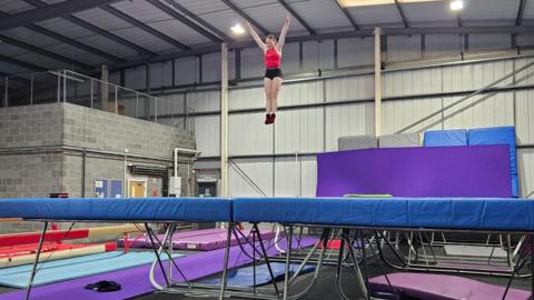 A large warehouse with multi-coloured mats on the floor and a trampoline  in the foreground. A girl is in the air above the trampoline 