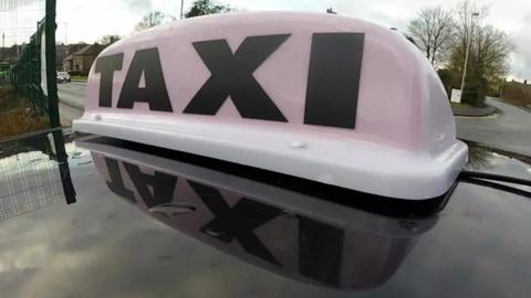 A close up generic image of a white taxi light with black writing which says TAXI on top of the roof a black car