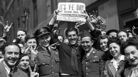 Crowds celebrate VE Day, holding up a newspaper that reads 'IT'S V-E DAY'