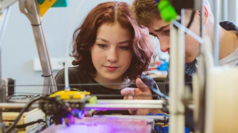 A teenage girl and boy looking at a 3D printer