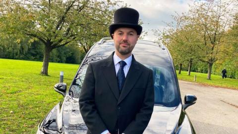 The image shows a funeral director standing in front of a hearse