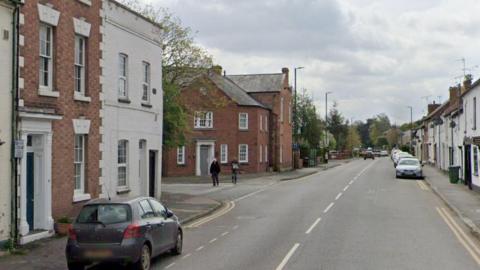 A Google view of the B4361 near Bargates. The residential area has cars parked in residential bays near terraced houses. Parts of the road have double yellow lines. People are walking on a pavement. There are trees in the background.