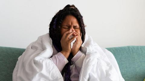 Young woman sitting on the sofa at home, covered with a blanket, blowing her nose, suffering from a cold or flu.