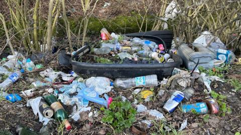 A wooded area where the muddy ground is covered in litter, including glass and plastic bottles, plastic bags, cans, tins and what appears to be a black plastic bin, which is partially buried and also covered with litter. Bare shrubs are in the background.