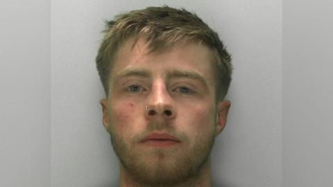 A mugshot of a man in his early 20s. He has shorter dirty blond hair and blond facial hair. He has a nose ring on one nostril. He has a blank expression and is standing in front of a plain grey background.
