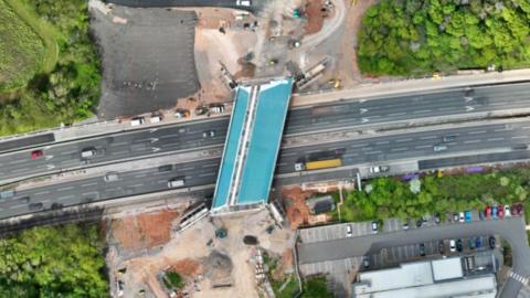 Image from a drone showing a motorway with a number of cars and lorries driving along it, a car park and building works. In the centre of the image is a new bridge under construction.