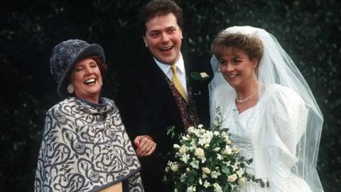 A woman wearing a grey hat and grey and cream shawl, stood next to a man wearing a black suit, white shirt and yellow tie, stood next to a woman dressed in a wedding veil and dress holding a bouquet of white flowers on their wedding day. All of them are smiling and looking happy.