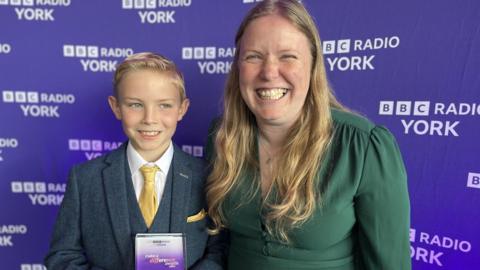 A young boy stands next to his mum. The boy has blue eyes, blonde hair and is wearing a smart blue-green tweed suit with a waistcoat and yellow tie. He is smiling and holding a glass award with 'Make A Difference Awards 2025' on it. His mum is wearing a green dress and has long blonde hair and is wearing a silver pendant necklace. She is also smiling. Behind them is a purple backdrop with 'BBC Radio York' on it. 