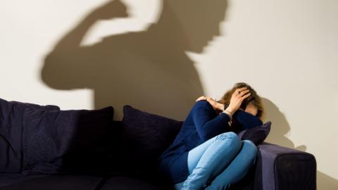 A woman cowers on a sofa while the shadow of a man with a clenched fist is clear on the wall behind her