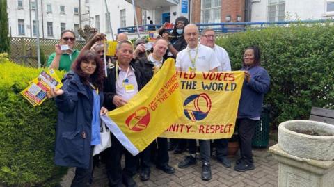 About a dozen people stood beside some bushes at the entrance to a building. They are carrying yellow leaflets and the people at the front of the group are carrying yellow flags bearing a megaphone icon and the words 'United Voices of the World'.