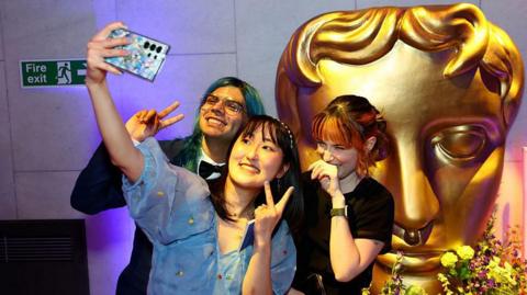 A group of three young people pose for a selfie in front of a large, golden Bafta mask statue. The young woman at the front of the group holds up her camera phone and throws up a peace sign, as does the young man with green hair to her right. A young woman to her left places the back of her hand against her mouth as she giggles.