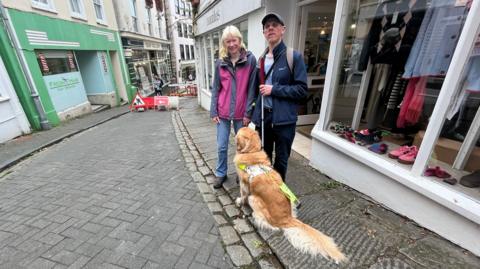 A man and his wife standing with a dog looking up at them sat wearing a harness, they are standing on a high street cobbled road with shops around it.