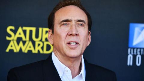 A close-up of Nicolas Cage seen with dark hair wearing a black jacket and white shirt, photographed at a red carpet event with a dark backdrop and yellow and blue logos behind him.