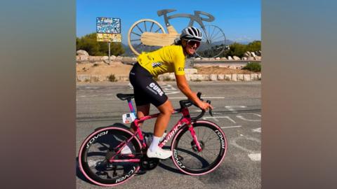 Michelle Sharland during the Tour de France Femmes. Michelle is wearing white cleets, black shorts and a yellow cycling top. She is on a pink bike with black wheels. She is wearing a silver helmet and is cycling in front of a giant silver bike statue.