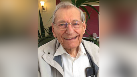 Mike Gibson smiles widely at the camera. He has thin grey hair, wears glasses and a light grey coat with a white shirt underneath. 
