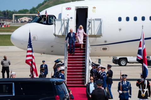 Plane shown on runway with King and Queen walking down the steps. There is a red carpet coming from the steps and military personnel can be seen carrying a US and Union flag. 