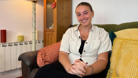 Antonia smiles at the camera as she sits on a sofa with a lamp next to her. Her brown hair is tied bag and she is wearing a beige shirt with her hands clasped together in the foreground.