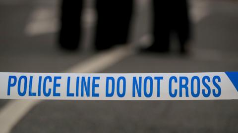 A 'police line do not cross' tape stretched across a road. The background is blurred.