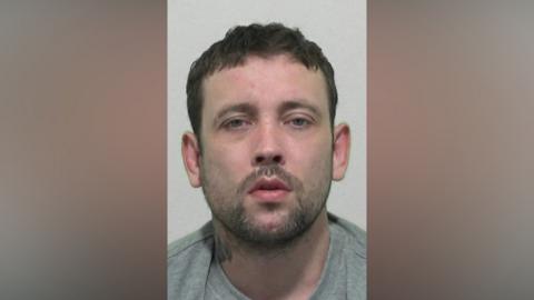 Mugshot of Wilkinson. He has a short dark beard and short black hair, with a tattoo on his neck.