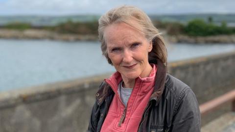 A woman with greying blonde hair in a dark jacket with a pink fleece underneath stands in front of Stranraer harbour