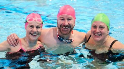 Two women and a man standing and smiling in a swimming pool. The women are wearing pink and green swimming caps and black swimming costumes. The man is wearing a cap and has goggles around his neck