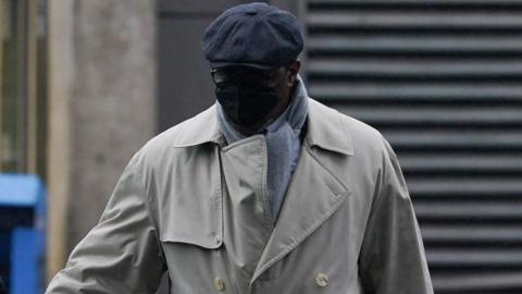 Charles Ehikioya wearing a beige trench coat, grey scarf, glasses, a black mask covering his face and a grey hat.