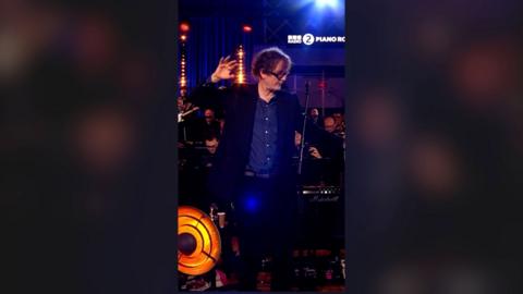 A man, Jarvis Cocker, in a blue shirt and suit, with one hand raised and outstretched, looking to the side.