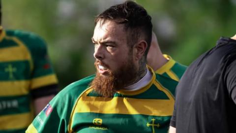 Connor Parker, wearing a yellow and green hooped rugby shirt, pictured during a match. He has a ginger beard and short hair. He is wearing a gumshield. 
