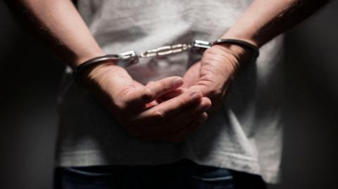 A stock image of a prisoner in handcuffs - the man wears a grey t-shirt and blue jeans and in pictured with his hands behind his back. The image is blurry.