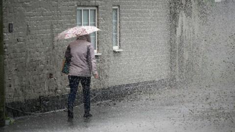 A woman walking away from the camera in heavy rain. She is wearing a grey jacket, with a bag over her left shoulder. She is carrying a floral umbrella. 