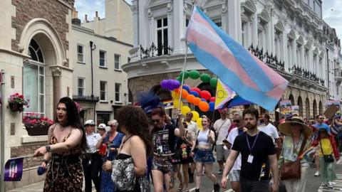 The photo shows a vibrant parade on a city street with participants in colourful attire, some holding balloons and flags. One person prominently carries a large transgender Pride flag. The background includes historic buildings with ornate architecture, flower boxes on the windowsills, and a blue sky.