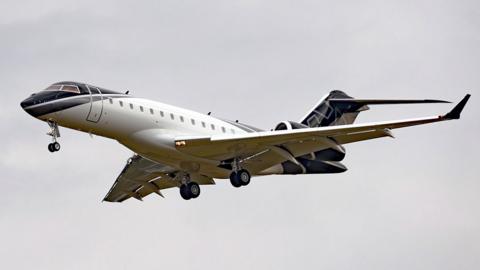 A generic image of a private jet against a grey sky, coming in to land