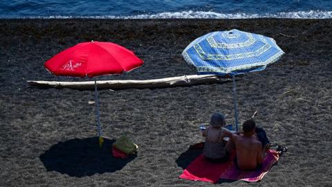 A red and blue parasol on a beach next to the sea, which is at the top of the picture. Two beachgoers are resting in the shade under the blue parasol on the right.