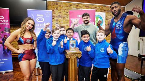 Six young boys in blue fleeces smile with their thumbs up. To the side of them are two muscular athletes. 