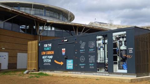 The new air source heat pump housed in a blue metal unit with pipes on display through windows and graphics on the side explaining how it works 