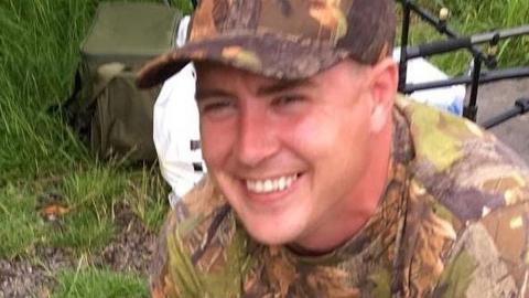 Paul Smith wearing a camouflage patterned t-shirt and cap with grass and a olive green bag in the background. He is smiling. 