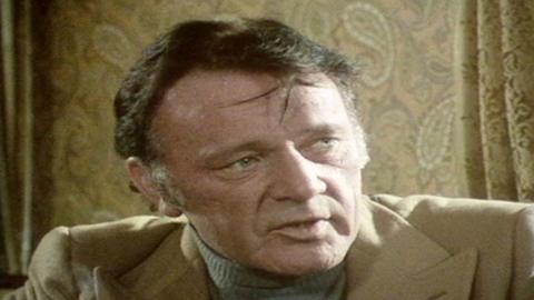 Richard Burton, with dark hair and grey sideburns, in a grey roll neck jumper and sandy coloured coat, looking off camera. There is beige wallpaper and curtains behind him 