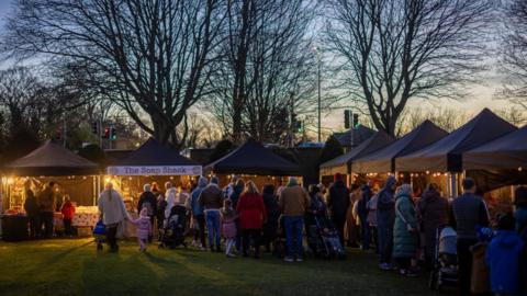 Crowds of people look at Christmas market stalls on a grassy lawn at twilight. 