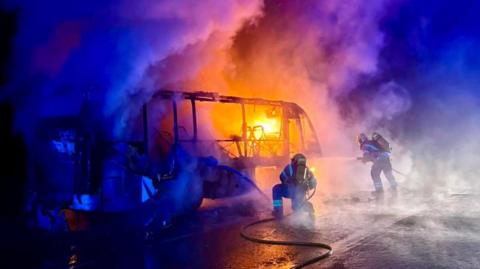 Firefighters with hoses spraying water on a bus on fire.